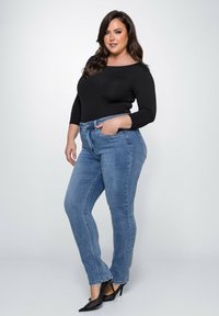 Schwarzes Langarmtop, blaue hochgeschnittene Skinny-Jeans und schwarze spitze High Heels. Glatte Textur und figurbetontes Design.