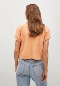 Kurzärmeliger, cropped Pullover aus strukturiertem orangefarbenem Strick. Hat eine lockere Passform mit umgeschlagenen Ärmeln. Wird mit hellblauen, hoch taillierten Jeans getragen.