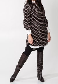 Czarna tunika z kwiatowym wzorem, długimi bufkami, białym kołnierzykiem i bocznymi rozcięciami, zestawiona z czarnymi rajstopami i brązowymi, wysokimi na kolano botkami.