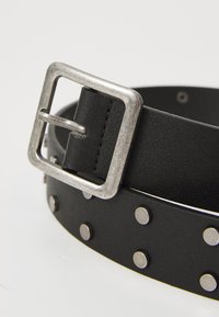 Curea neagră din piele, cu o cataramă metalică argintie și cu cuie decorative argintii pe toată lungimea. Textură netedă și design durabil.