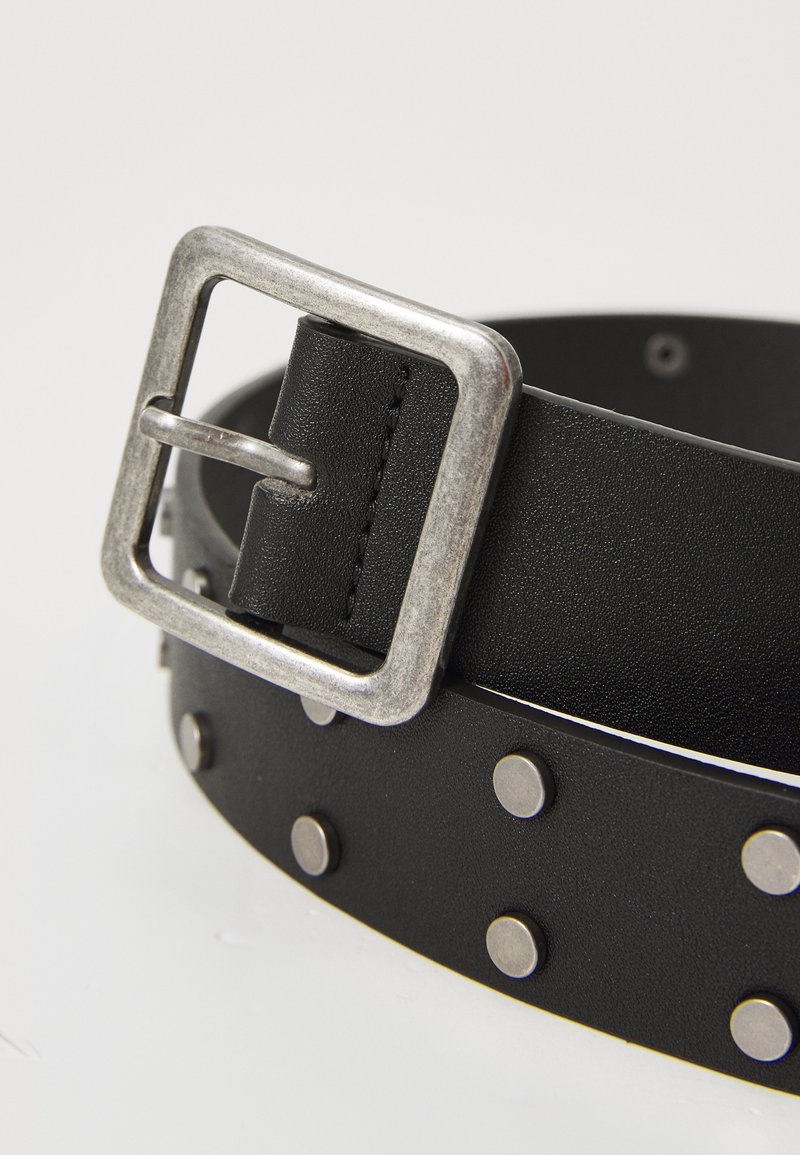 Cinturón de cuero negro con una hebilla de metal plateado y tachuelas decorativas de plata a lo largo de su longitud. Textura suave y diseño duradero.