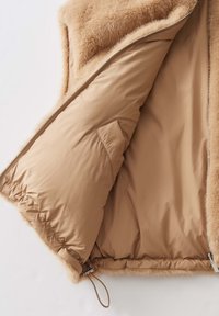 Beige Weste mit Kunstfell-Innenfutter und glattem Außenmaterial. Mit einem Frontreißverschluss und einem elastischen Saum. Die Innentasche ist sichtbar.