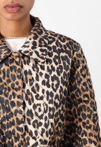 Gros plan sur une personne portant une veste matelassée à imprimé léopard avec un col, par-dessus un haut blanc texturé, sur un fond uni.