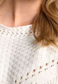 Hvid strikket sweater med et tekstureret mønster, der har en løs halsudskæring og små dekorative huller i hele stoffet.