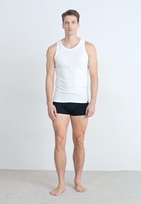 Un débardeur en coton blanc et un short ajusté noir sur un modèle masculin debout, pieds nus, contre un fond blanc uni.