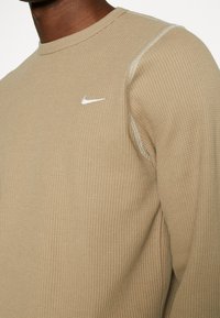 Chemise beige en maille gaufrée à manches longues, dotée d'un col rond et d'un petit logo Nike blanc sur la poitrine, avec des coutures contrastantes.