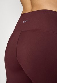 Burgundin venyvät housut, joissa on sileä, teksturoitu kangas. Ominaisuuksiin kuuluu korkea vyötärö ja pieni hopeinen Nike-logo takana.