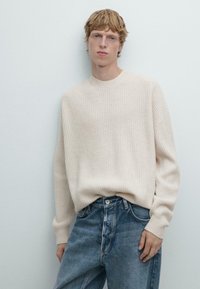 Giovane con capelli biondi corti, vestito con un maglione cremoso a coste e jeans blu larghi, in piedi contro un muro chiaro.