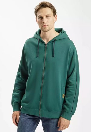 Zielona bluza z kapturem na zamek, wykonana z miękkiego materiału, z przednim zamkiem, kapturem ze sznurkiem i bocznymi kieszeniami. Posiada kontrastowe przeszycia oraz ściągacze przy mankietach.