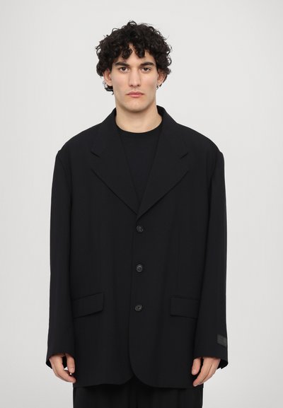 Černý oversized blazer s klopovými límci, třemi knoflíky a předními kapsami. Hladká tkanina s minimálními detaily a volným střihem.