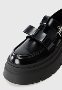 Mocassins à plateforme noirs avec une tige en cuir brillant, présentant un large nœud décoratif et une semelle en caoutchouc texturée.