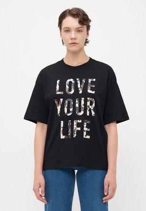Osoba s krátkými vlasy, oblečená v černém tričku s nápisem "LOVE YOUR LIFE" v béžovém a hnědém maskáčovém vzoru a modrých džínách.