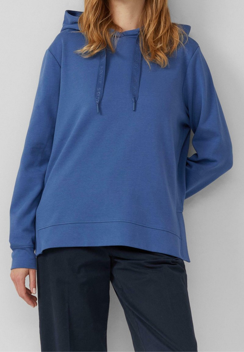Femme portant un sweat à capuche bleu avec des cordons et un pantalon sombre, debout devant un fond uni gris clair.