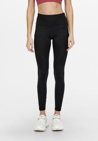 Leggings neri a vita alta con una texture liscia, caratterizzati da dettagli di cuciture curve e tasche laterali, abbinati a sneaker bianche.