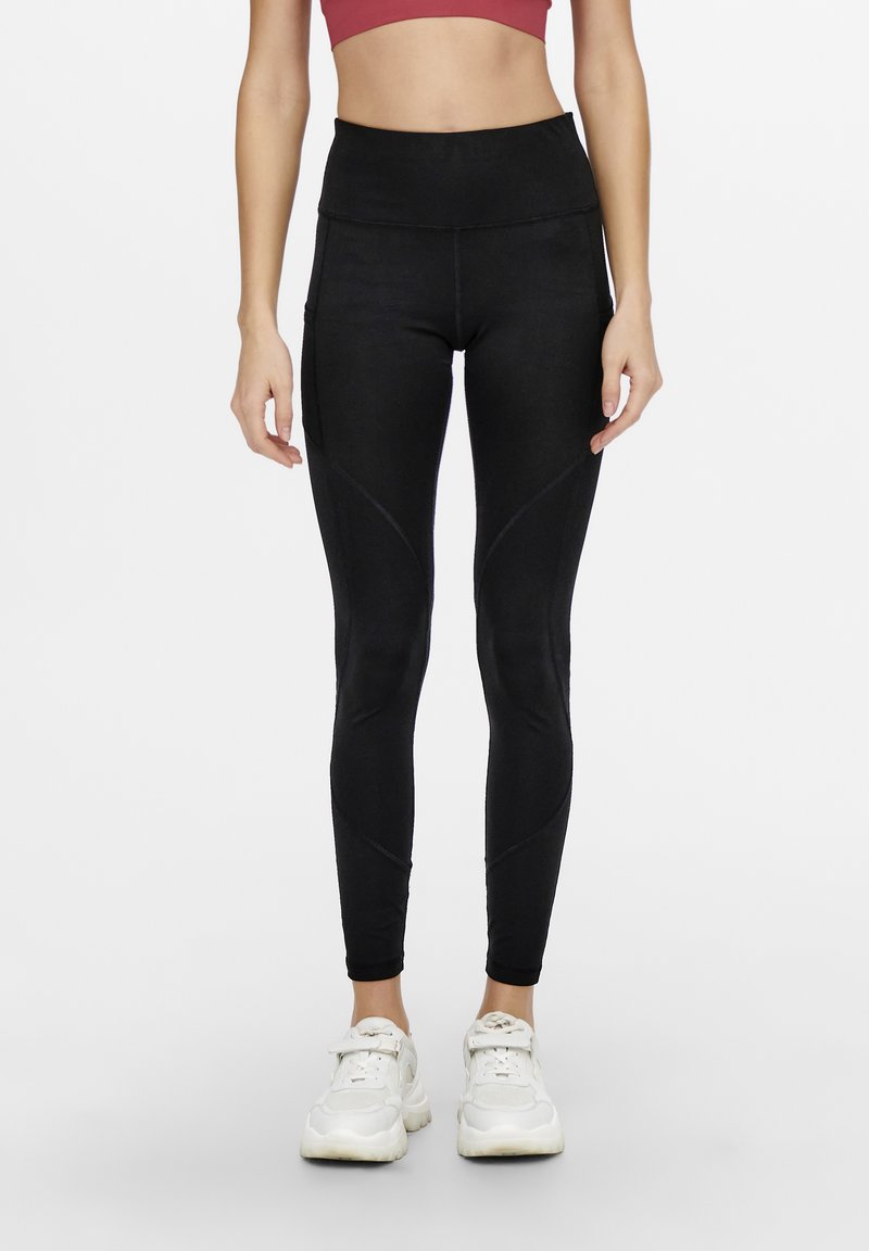 Leggings neri a vita alta con una texture liscia, caratterizzati da dettagli di cuciture curve e tasche laterali, abbinati a sneaker bianche.