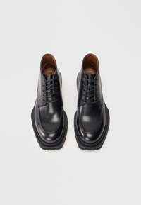 Botas de vestir de cuero negro con cordones, suelas gruesas, punteras cuadradas redondeadas y costuras visibles, vistas desde arriba sobre un fondo blanco.