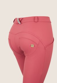 Pantaloni da equitazione rosa con vita alta, texture liscia, cuciture curve e un dettaglio decorativo dorato sulla tasca posteriore.