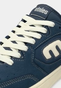 Blue suede sneaker with white laces and rubber sole. Features a logo on the side and contrasting stitching detail.