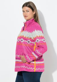 Rosa Fleece-Pullover mit einem Half-Zip, gemustert mit weißen, orangen und grauen Zickzacklinien, ausgestattet mit Seitentaschen und gelben Reißverschlussakzenten.