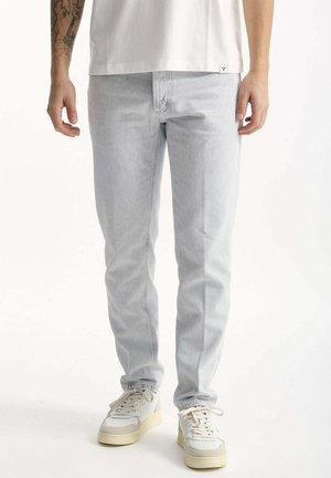 Uomo che indossa jeans aderenti azzurri chiaro, sneakers bianche e una maglietta bianca, in piedi davanti a uno sfondo bianco neutro.