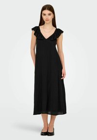 Una joven se encuentra de pie vestida con un vestido midi sin mangas negro con escote en V con volantes y bailarinas negras sobre un fondo claro.