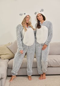 Due persone indossano pigiami morbidi a tema panda, grigi e bianchi, con cappucci che presentano orecchie e chiusura con zip, su uno sfondo di un divano chiaro.