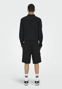 Zwarte lange mouwen shirt gecombineerd met zwarte cargoshorts, voorzien van zijzakken. Het model draagt witte sokken met zwarte strepen en zwarte sneakers.