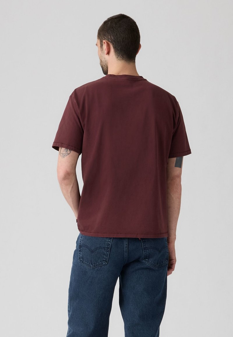 Uomo visto da dietro che indossa una maglietta bordeaux tinta unita e jeans blu su uno sfondo grigio chiaro uniforme.