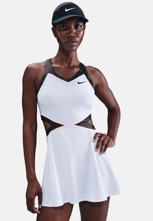 Athletische Frau, die ein weißes und schwarzes ärmelloses Nike Tenniskleid mit Aussparungen an der Taille und ein schwarzes Nike-Visier trägt.