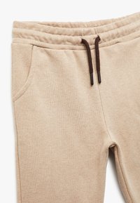 Beige sweatpants lavet af blødt materiale med en elastisk talje, justerbare brune snore og sidelommer. Tekstureret stofdetalje.