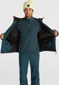Veste d'extérieur teal ouverte sur un haut en polaire teal foncé et un pantalon assorti. Un bonnet jaune clair repose sur la tête de la personne.
