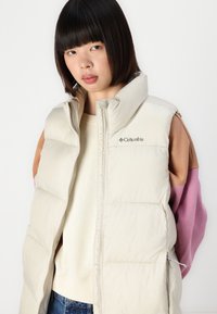 Beige puffer vest met een hoge kraag, voorzien van een rits aan de voorkant en het Columbia-logo. Draagt over een lichte sweater en een kleurrijke longsleeve.