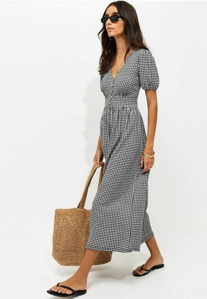 Kobieta ubrana w czarno-białą sukienkę midi w kratę gingham z bufiastymi rękawami, czarne okulary przeciwsłoneczne, sandały, niesie dużą plecioną torbę typu tote.