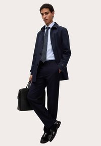 Marineblauer Trenchcoat über einem blauen Hemd mit Streifen-Krawatte, kombiniert mit Nadelstreifenhosen und glänzenden schwarzen Schuhen, trägt eine schwarze Tasche.