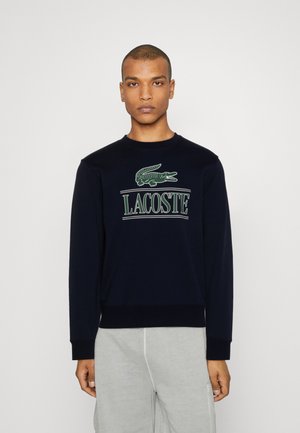 Sweat-shirt bleu marine en mélange de coton, mettant en avant un logo de crocodile vert et le texte « LACOSTE » en blanc. Poignets et ourlet côtelés.