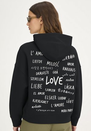 Frau trägt schwarzen Hoodie mit dem Wort "love", das in mehreren Sprachen auf dem Rücken gedruckt ist, stehend vor einem einfarbigen Hintergrund.