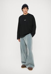 Juodas ilgomis rankovėmis marškinėliai su kontrastinga siūlė, šviesiai mėlynos plačios džinsinės kelnės ir rudos šlepetės su subtiliais akcentais.