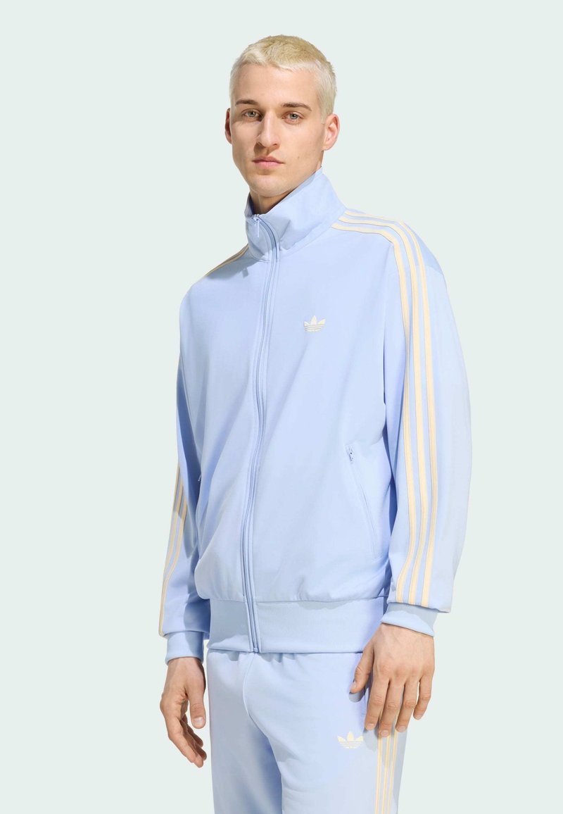 Young man with short blond hair wearing a light blue Adidas tracksuit with pale yellow stripes, standing against a plain background.