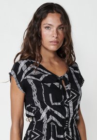 Zwarte jurk met witte geometrische patronen, korte mouwen, een strik bij de halslijn en een getailleerde taille, gemaakt van lichtgewicht stof.
