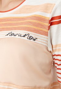 Top a rayas en tejido suave con tonos naranja, melocotón y crema. Presenta un escote redondeado y un detalle bordado que dice "Ahora es Ahora".
