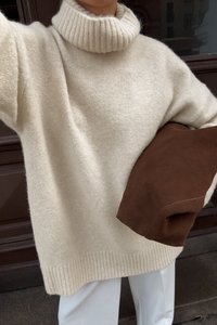 Cremfarbener, grob gestrickter Pullover mit einem gerippten Rollkragen und Saum. Kombiniert mit einer braunen Wildleder-Clutch, die eine weiche Textur zeigt.
