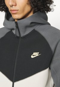 Felpa con zip caratterizzata da blocchi di colore nero, grigio e crema. Il tessuto è morbido e presenta cuciture visibili, con un logo Nike dorato sul petto.