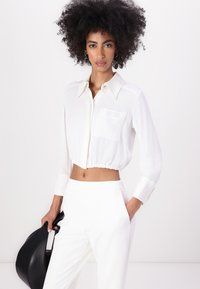 Bílé zkrácené blouse s límcem na knoflíky, elastickým lemem a kapsou. V kombinaci s bílými kalhotami a černou kabelkou. Hladká tkanina.