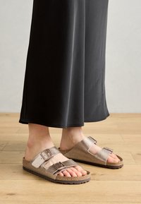 Sandați slip-on din piele maro, cu două curele ajustabile și catarame metalice, pe o talpă de plută, asortați cu pantaloni negri cu picioare largi.