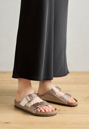 Braune Leder-Sandalen zum Hineinschlüpfen mit zwei verstellbaren Riemen und metallischen Schnallen, auf einer Korksohle, kombiniert mit schwarzer, weit geschnittener Hose.