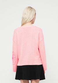 Ροζ πλεκτό πουλόβερ με χαλαρή εφαρμογή, με χαμηλωμένους ώμους και σχέδιο λουλουδιού σε ροζ στο αριστερό μανίκι. Ριμπ τελείωμα και μανσέτες.