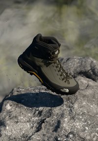 Buty wędrówkowe w ciemnooliwkowym kolorze, z chropowatą cholewką, żółtymi akcentami i wytrzymałą podeszwą. Widoczne są detalowe przeszycia i sznurowadła.