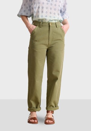 Olivgrüne hochgeschnittene Hose mit lockerer Passform, seitlichen Taschen, umgeschlagenen Säumen und flachem Design. Kombination mit braunen Sandalen.