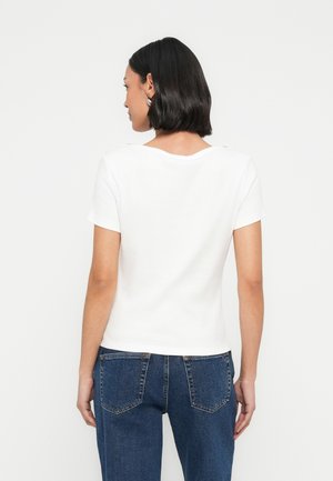 Vrouw met kort zwart haar, gekleed in een wit topje met korte mouwen en blauwe spijkerbroek, van achteren tegen een effen witte achtergrond.