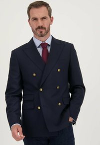 Marineblauwe dubbelzijdige blazer met gouden knopen, gestructureerde schouders en twee voorkezen. Gecombineerd met een lichtblauw shirt en een bordeauxrode stropdas.